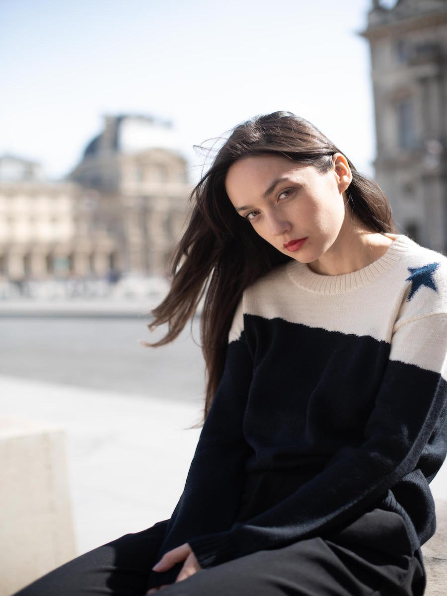 A black and cream alpaca sweater with a blue star detail on the shoulder, worn by a model in an outdoor city setting