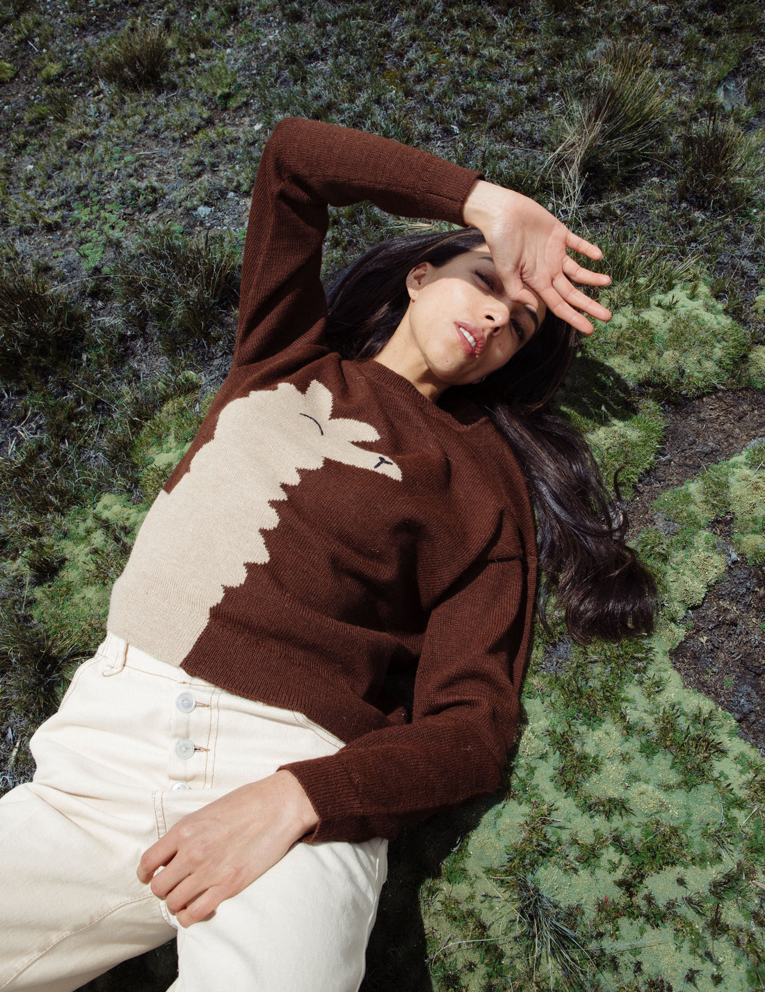 Woman wearing a brown sweater with a white design, sitting on grass.