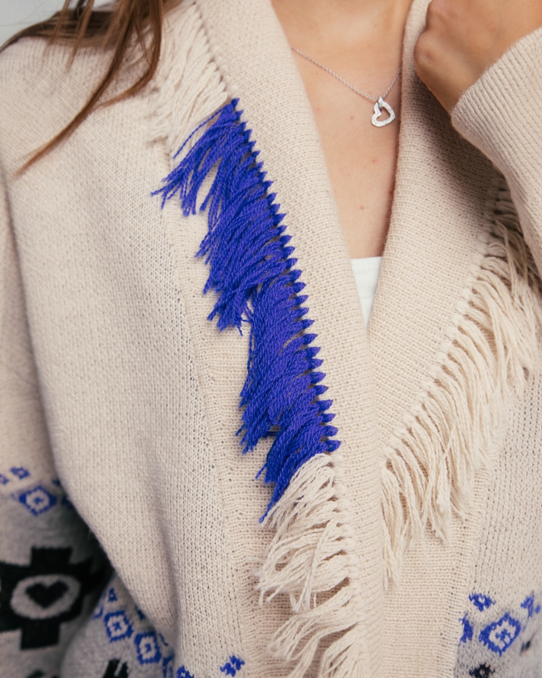 Close-up of a beige cardigan with blue fringe details and a heart-shaped necklace.