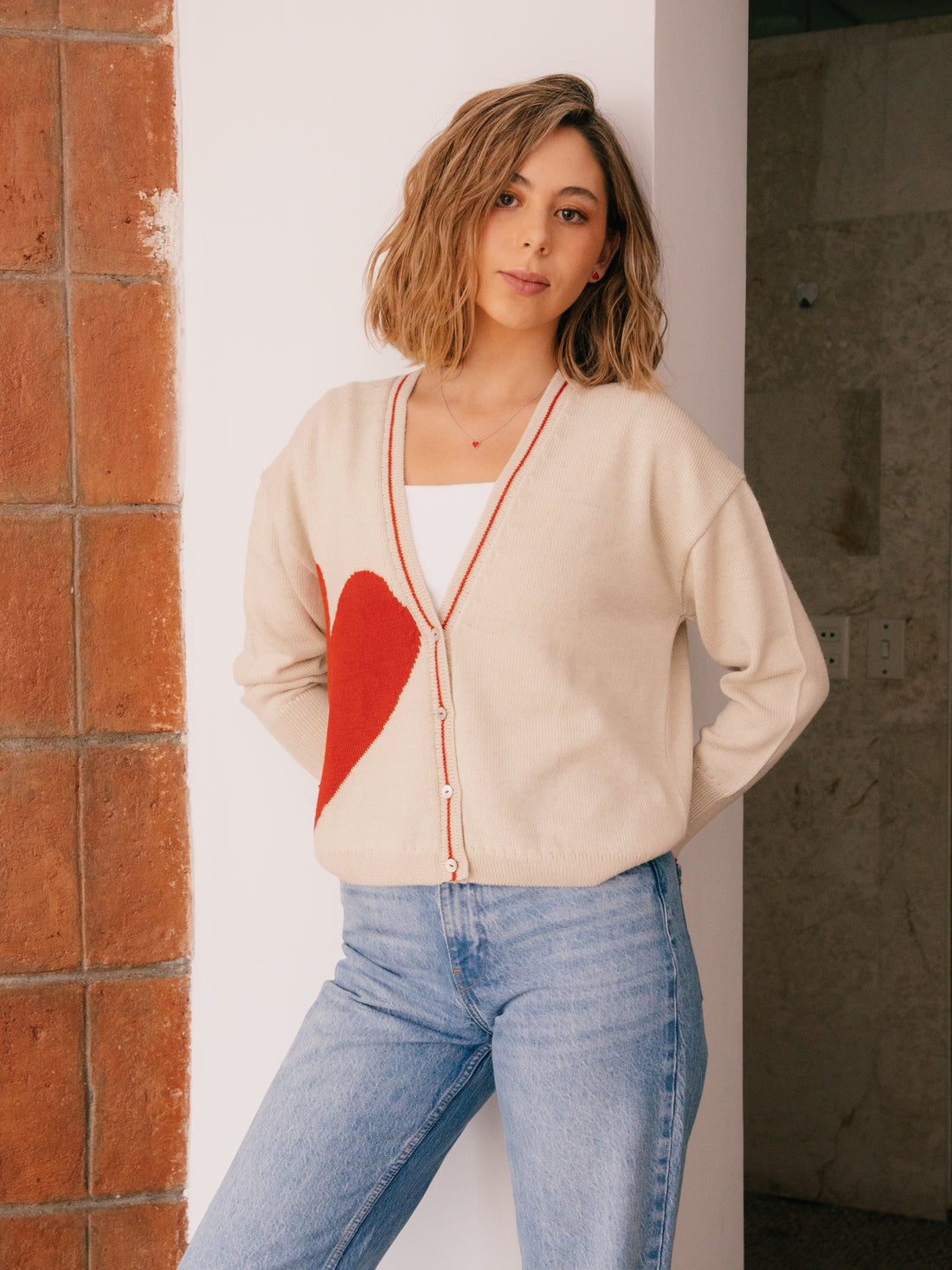 Woman wearing a beige cardigan with a red heart design, standing against a neutral background.