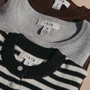 Three folded YAPA knit cardigans stacked on a beige background: a dark brown one on top, a light gray one in the middle, and a black-and-white striped one at the bottom.