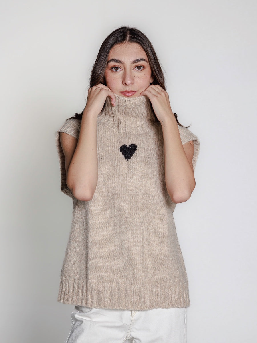 Woman wearing a beige knitted alpaca sweater vest with a black heart design on a plain background