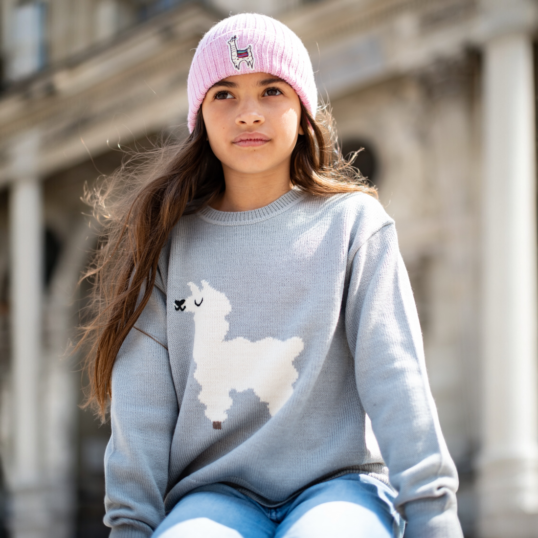 Girl wearing an alpaca pink beanie with a light blue sweater with a llama design beanie outdoors.