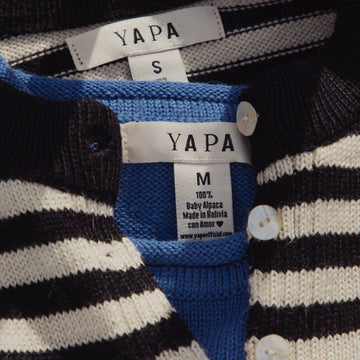 Close-up of a striped knit cardigan sweater with black and off-white horizontal stripes, featuring a blue inner layer and white buttons. The inner neck labels show the brand YAPA and size information.