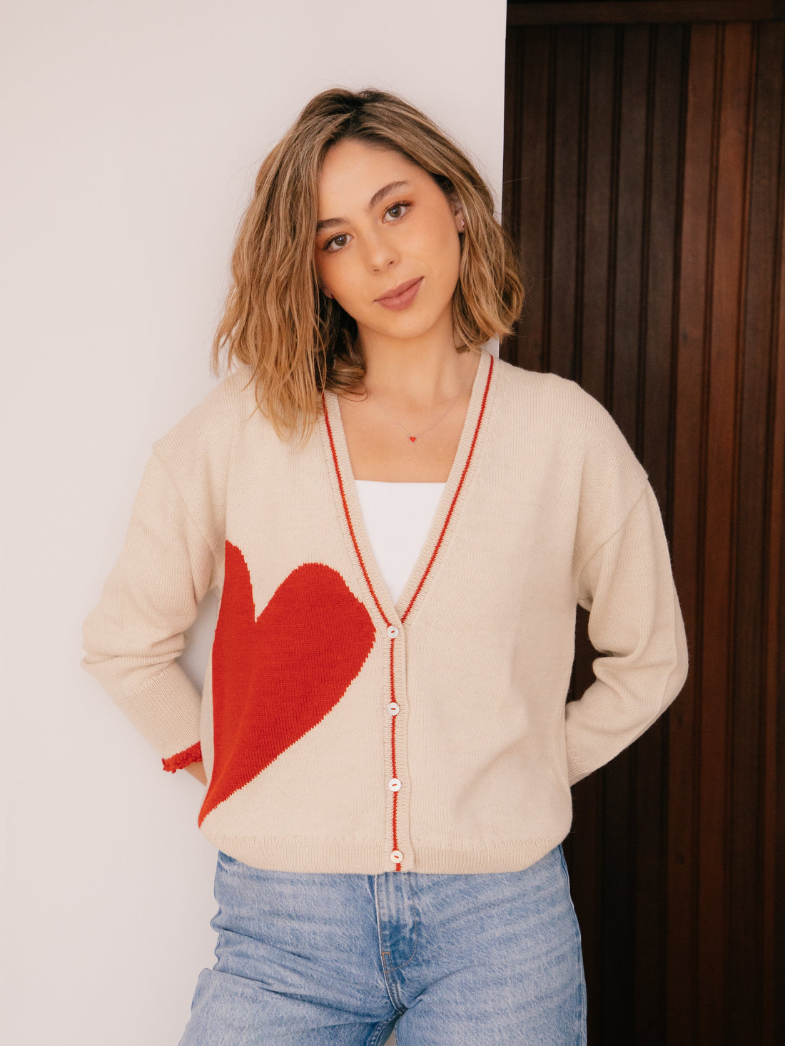 Woman wearing a beige cardigan with a red heart design, standing against a neutral background.