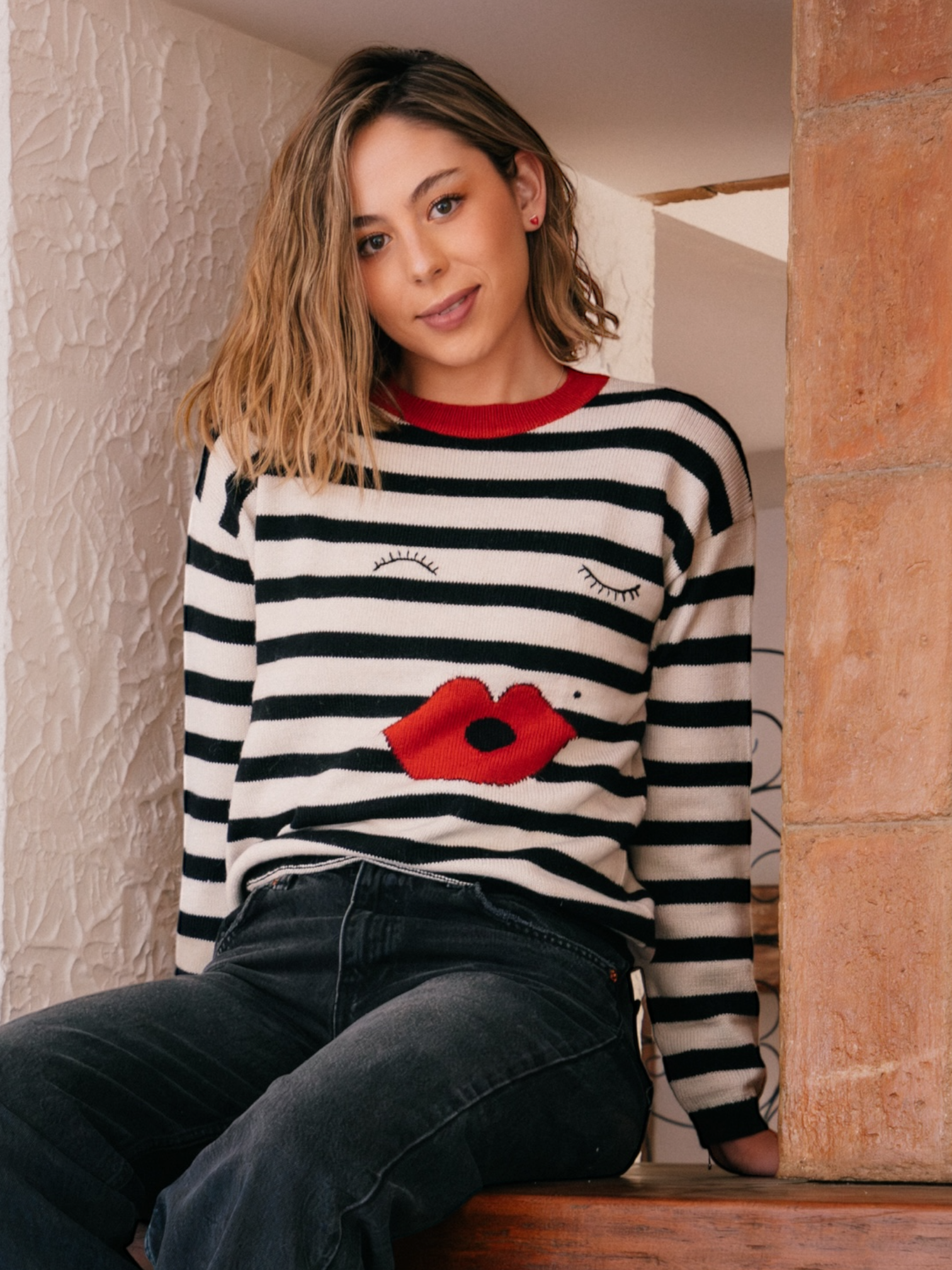 Woman wearing a black and white striped sweater with a red poppy design, sitting against a textured wall.