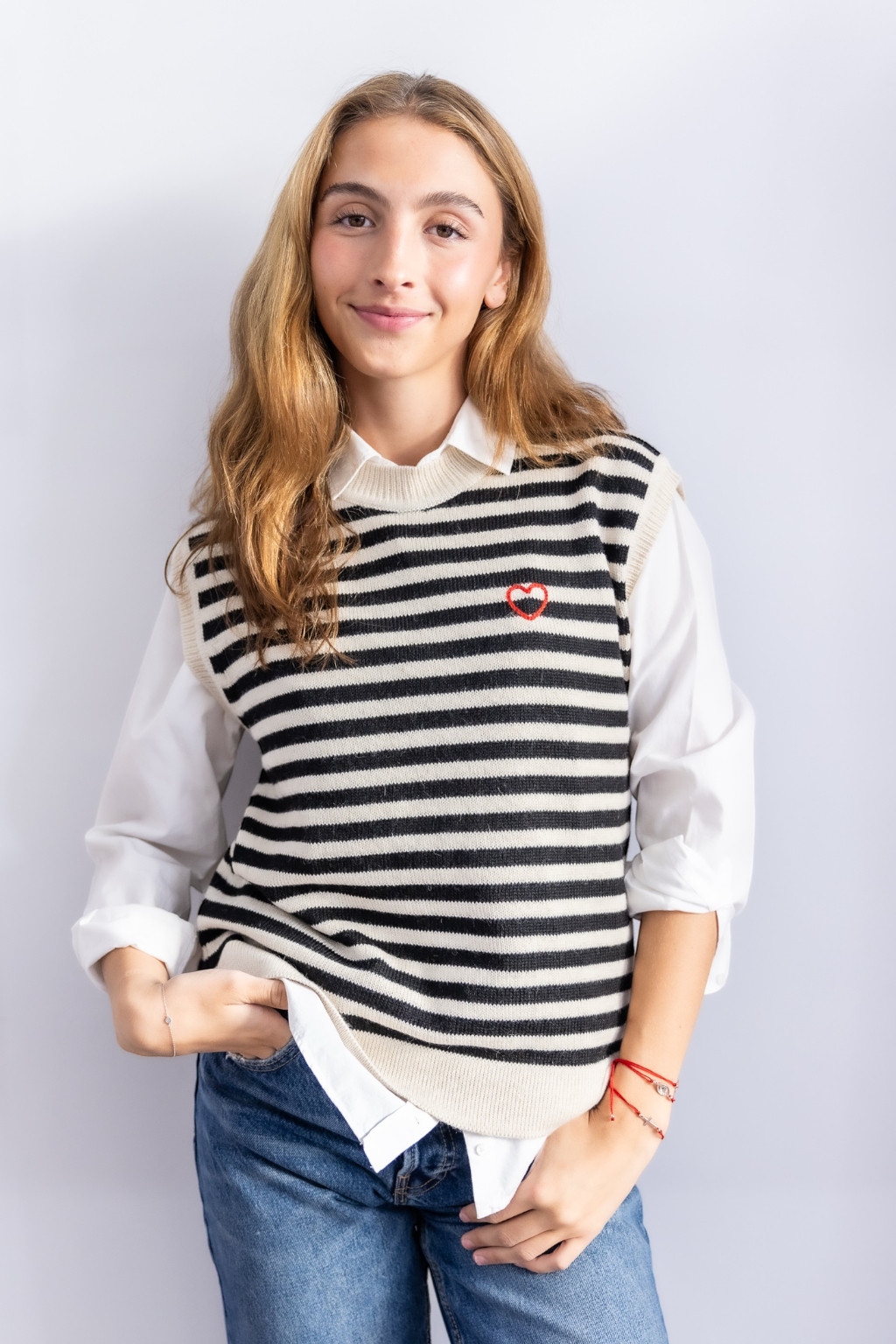 Person wearing a striped sweater vest with an embroidered heart design against a plain background
