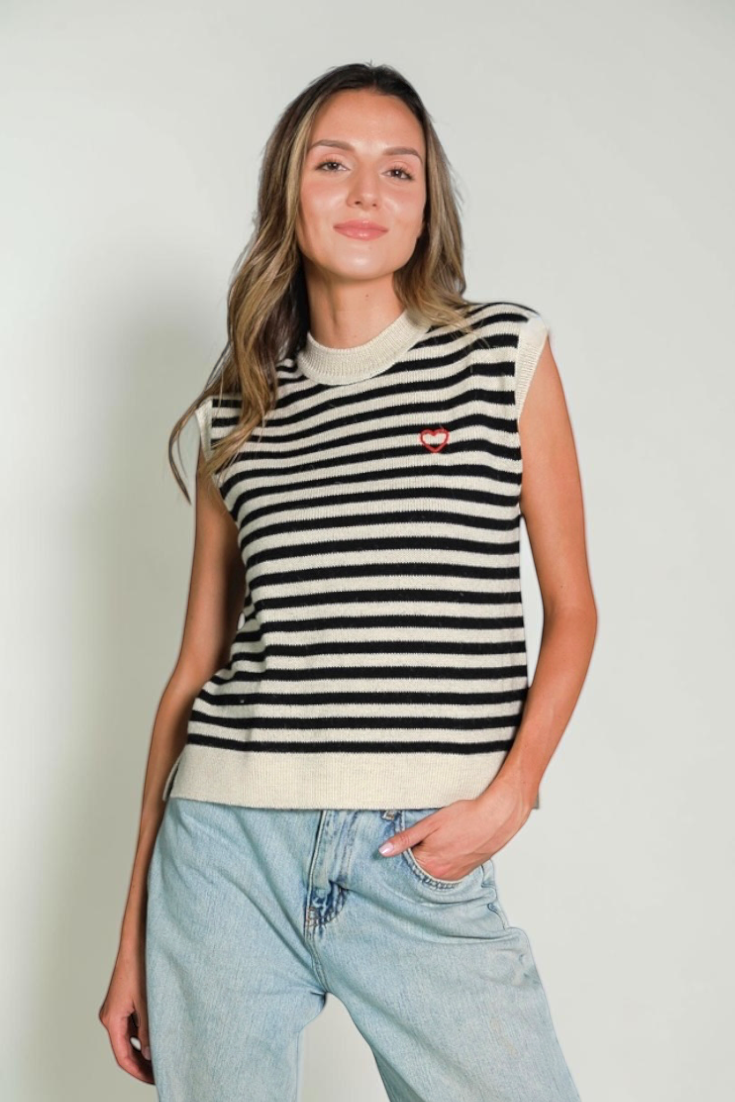 A woman modeling a sleeveless knit sweater vest with horizontal black and cream stripes, featuring a small red embroidered logo on the left chest, paired with light-wash blue jeans against a plain light gray background.