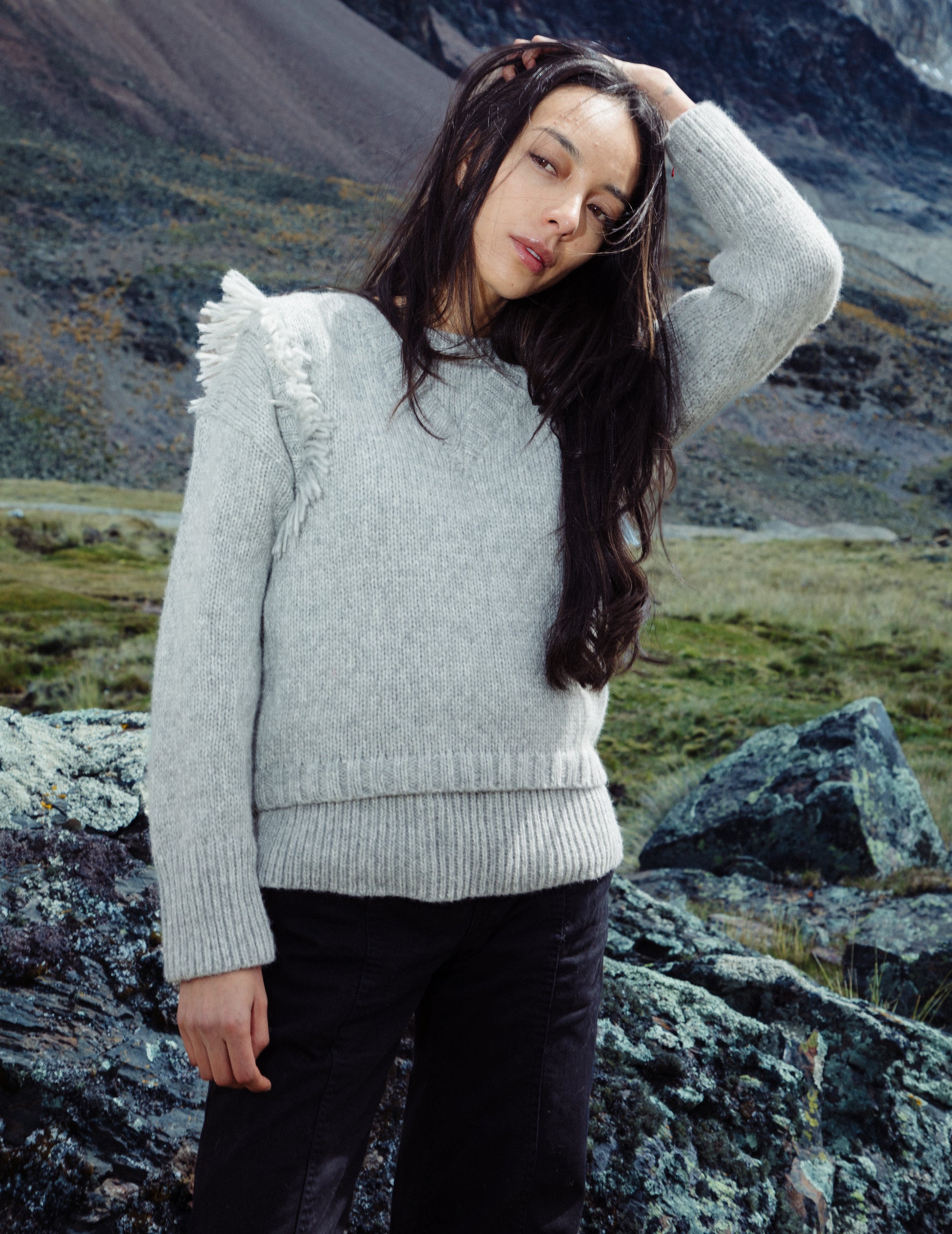 Woman in a light gray sweater standing on rocky terrain with mountains in the background