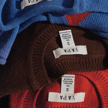 Three knitted crewneck sweaters stacked together, showing the inner neck labels. The top sweater is medium blue, the middle is dark brown, and the bottom is red-orange.