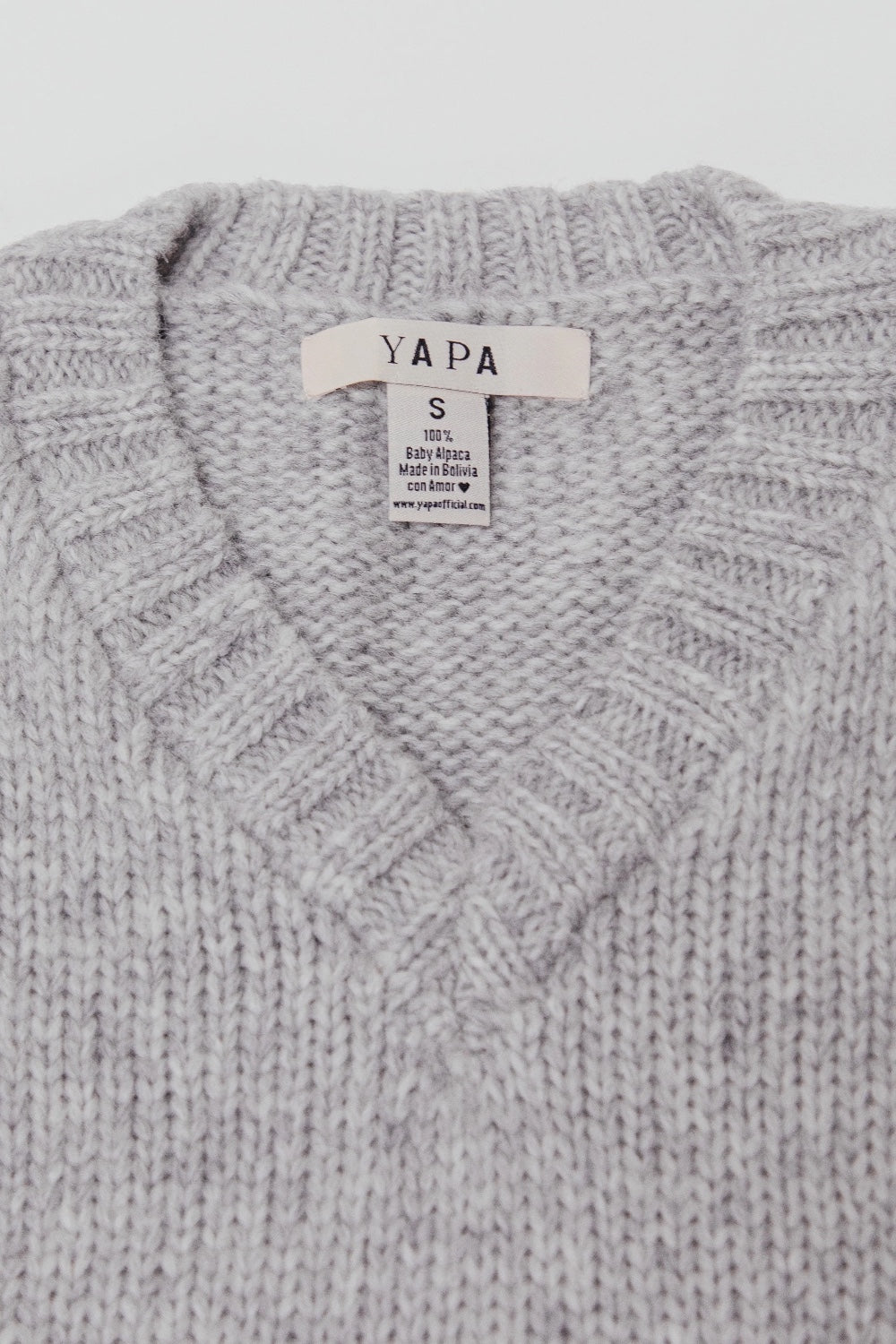 Close-up of a light gray knit sweater with a ribbed V-neck collar and a visible YAPA brand label.