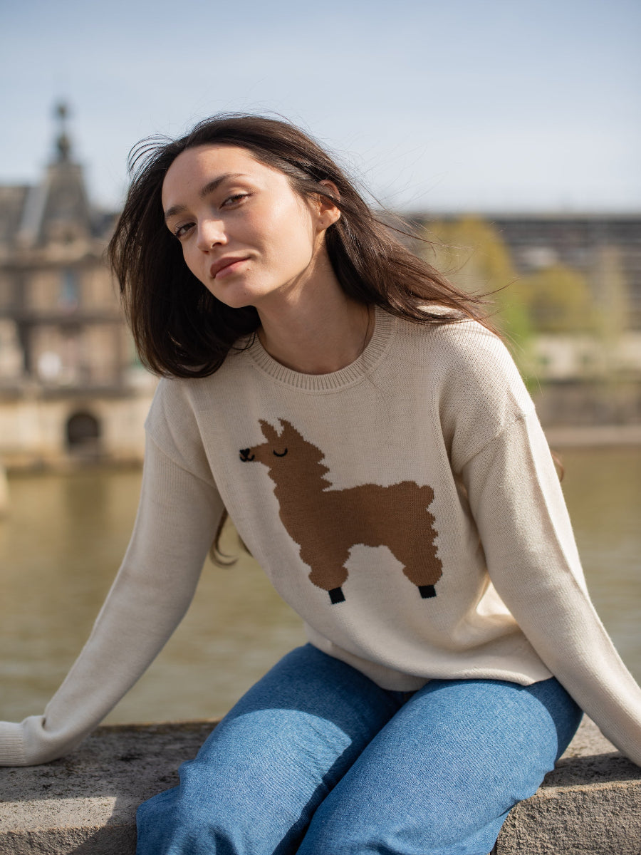 beige alpaca sweater with a brown llama motif, worn by a model styled with blue jeans with a city and river background.