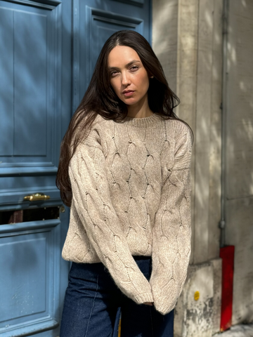 beige llama cable-knit sweater, worn by a model standing outdoors against a blue door