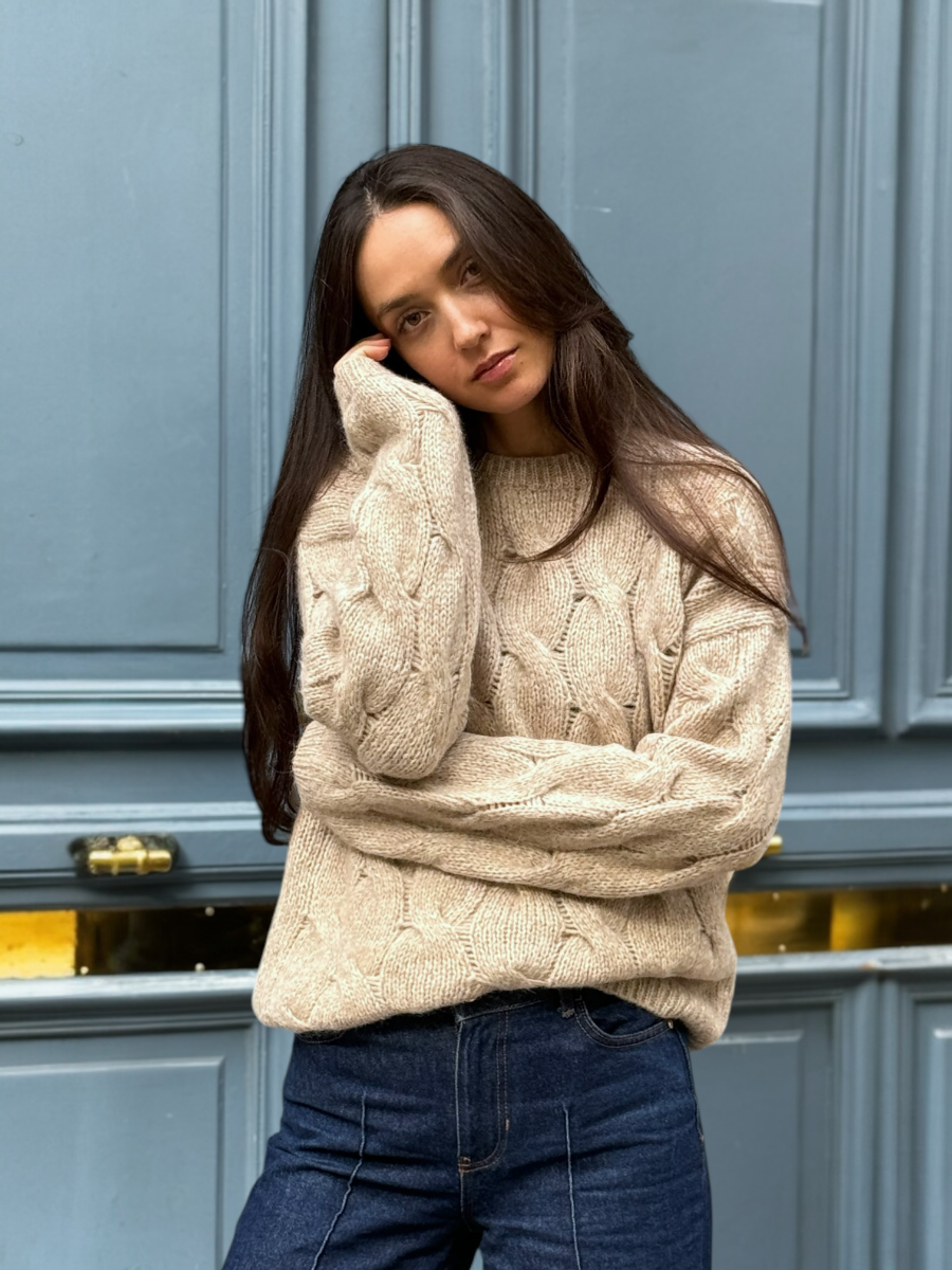 beige llama cable-knit sweater, worn by a model standing outdoors against a blue door