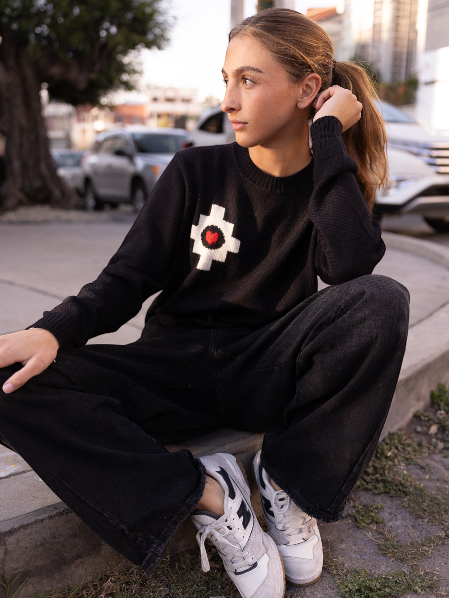 black alpaca sweater featuring a white cross pattern with a red heart at the center design, worn by a model sitting on the street