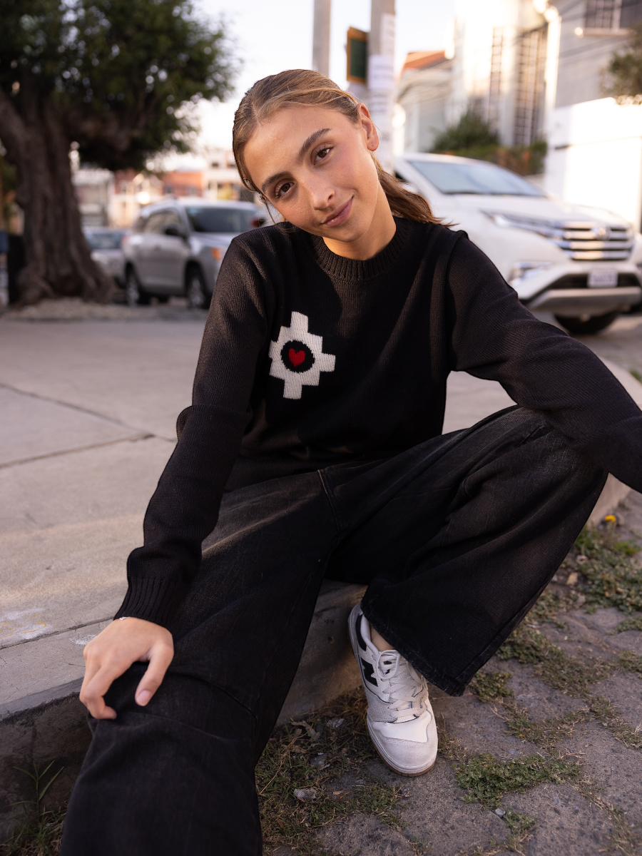black alpaca sweater featuring a white cross pattern with a red heart at the center design, worn by a model sitting on the street