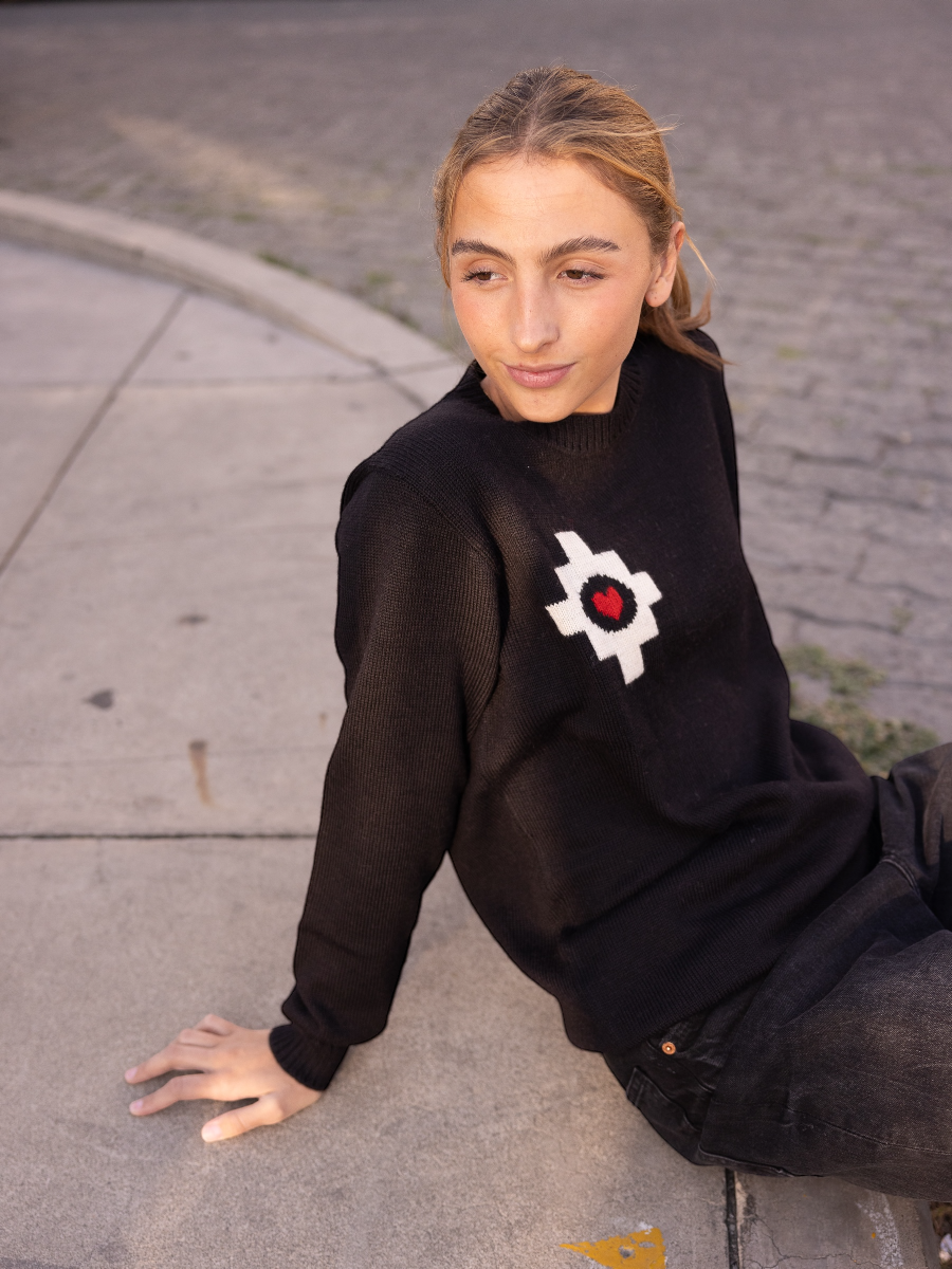 black alpaca sweater featuring a white cross pattern with a red heart at the center design, worn by a model sitting on the street