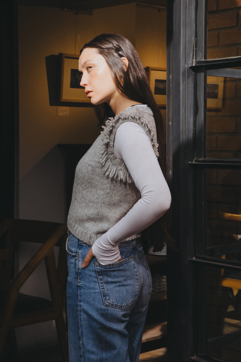 A woman posing in a gray knit sweater vest with frayed shoulder details over a white long-sleeve shirt and blue jeans, set against a dark textured wall.