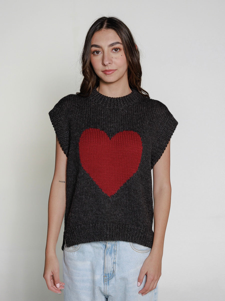 Woman wearing a grey knitted vest with a burgundy heart with blue jeans on a plain background