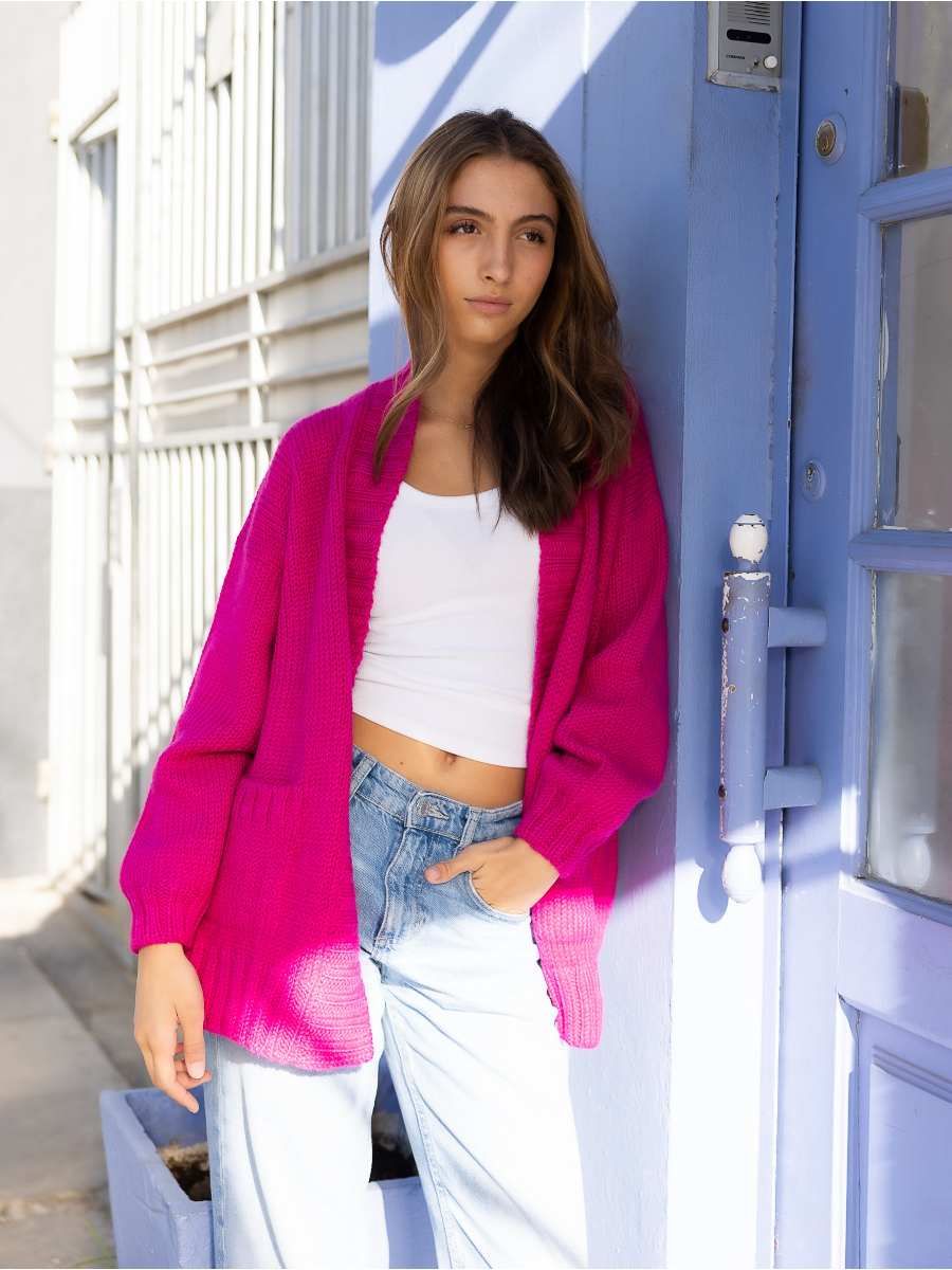 A woman wearing a pink knitted alpaca wool cardigan with a red heart on the back in a city street.