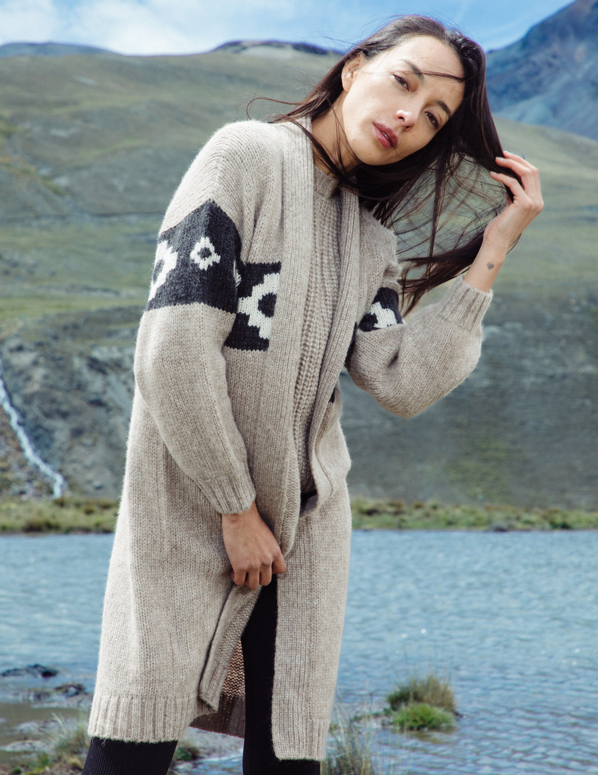 Woman wearing a long, patterned cardigan by a lake with mountains in the background