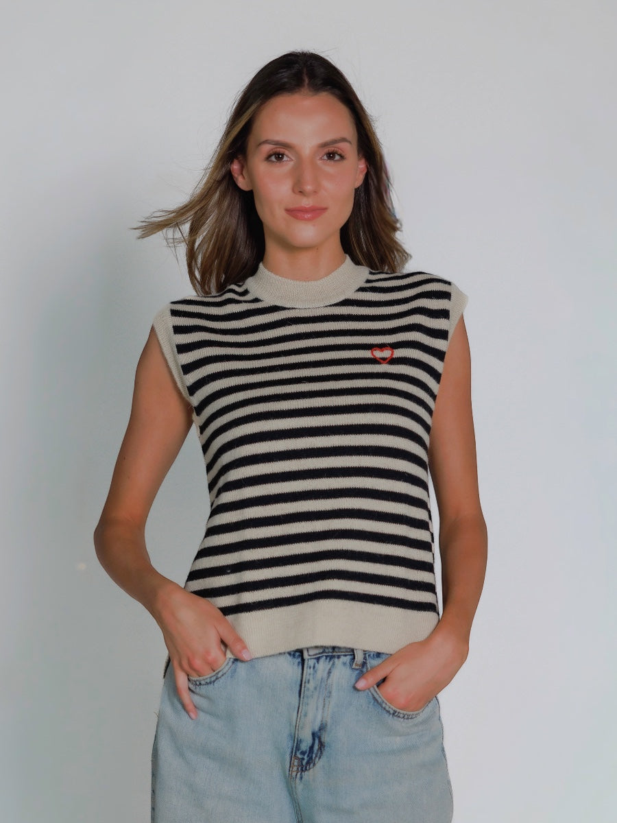 Woman wearing a black and white striped sleeveless alpaca vest with a hand embroidered heart and light blue jeans against a plain background
