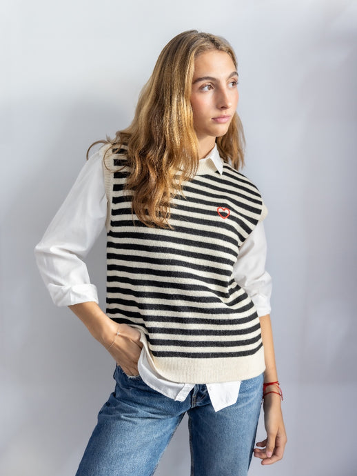 Woman wearing a black and white striped knitted vest over a white blouse with blue jeans on a plain background
