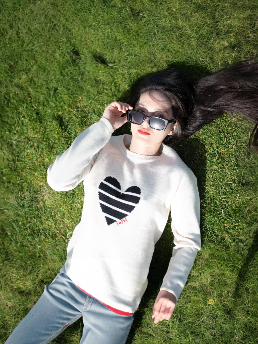 white alpaca knitted sweater with a black striped heart design, worn by a model lying on the grass