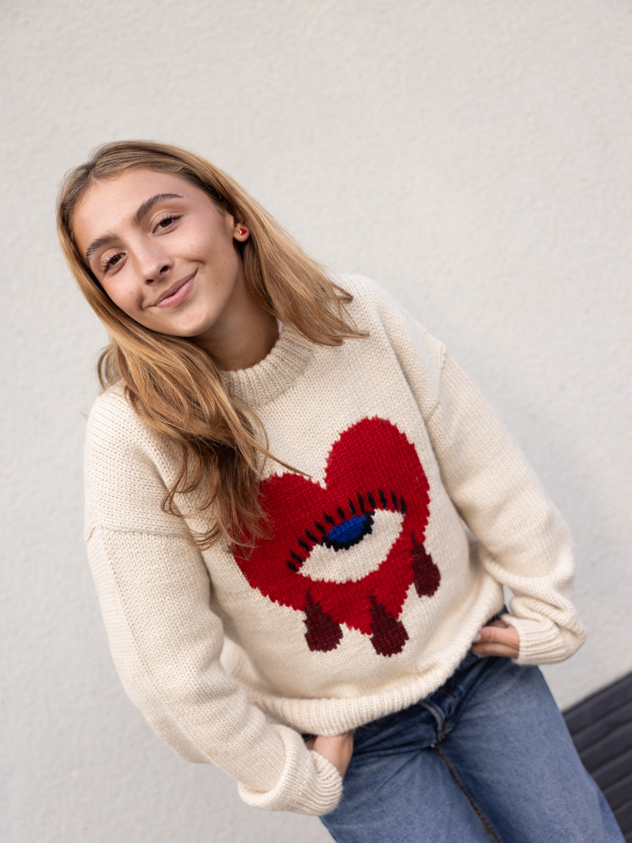 white alpaca sweater featuring a bold red heart motif with a blue eye and tear details on the front, shown here on a model against a plain background.