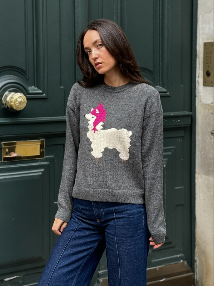 Woman wearing a grey sweater with a llama with a pink hat design, standing against a green door.