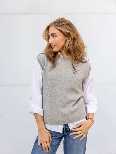 A woman wearing  a knitted vest with a round neckline, featuring a hand-embroidered heart on the chest.