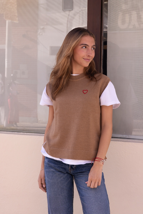 A woman wearing  a brown knitted vest with a round neckline, featuring a hand-embroidered heart on the chest.