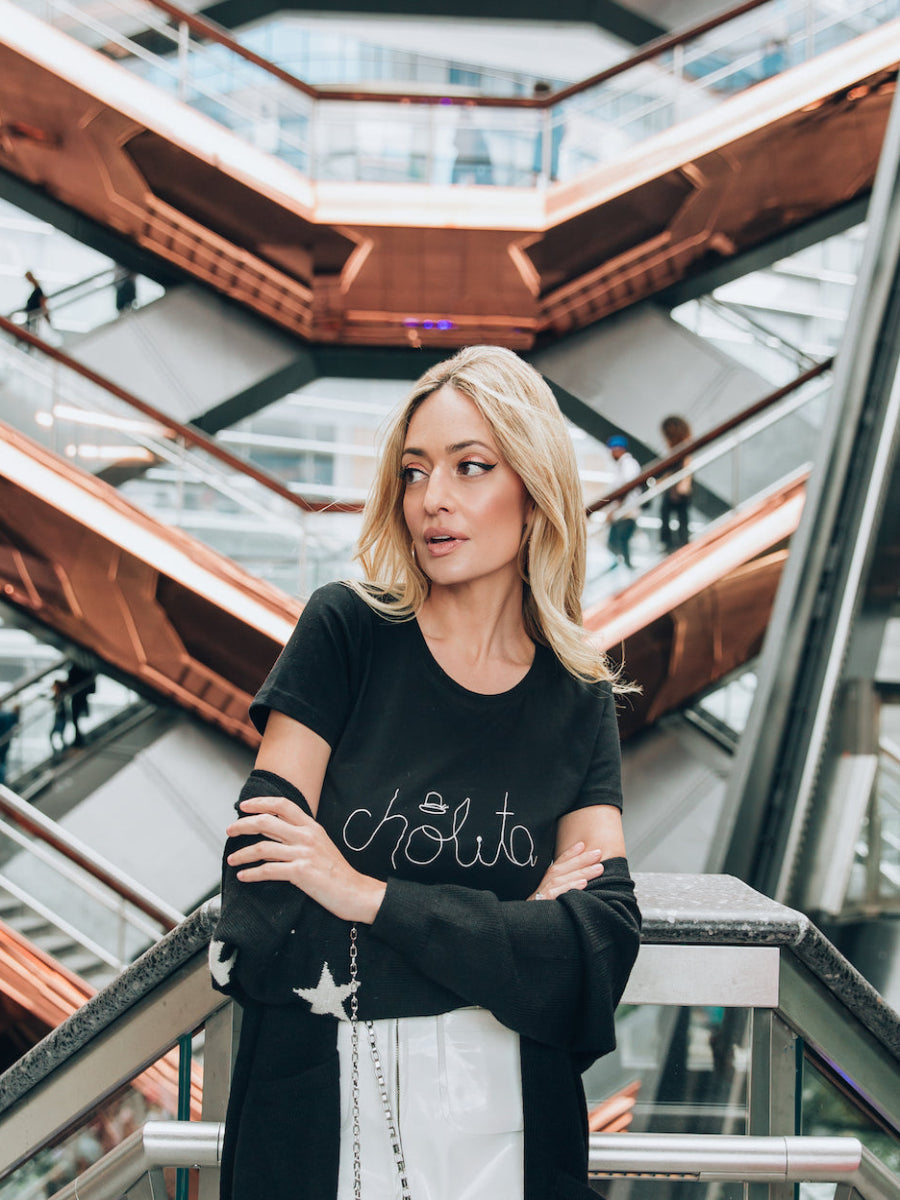 Black cotton t-shirt featuring the word “cholita” embroidered in white script on the front, shown here on a model in a modern urban setting.