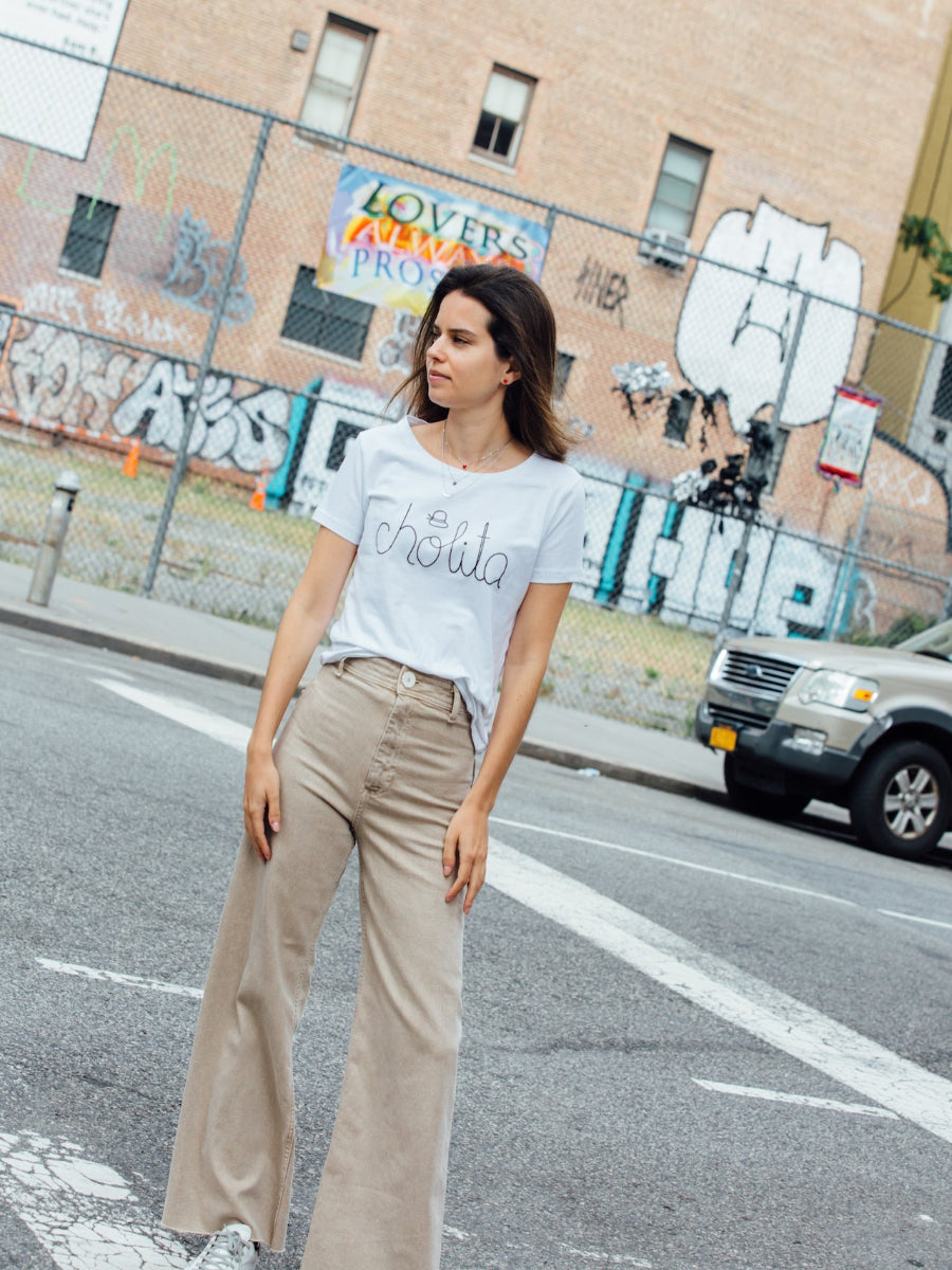 White cotton t-shirt featuring the word “cholita” embroidered in black script on the front, worn by a model in the street