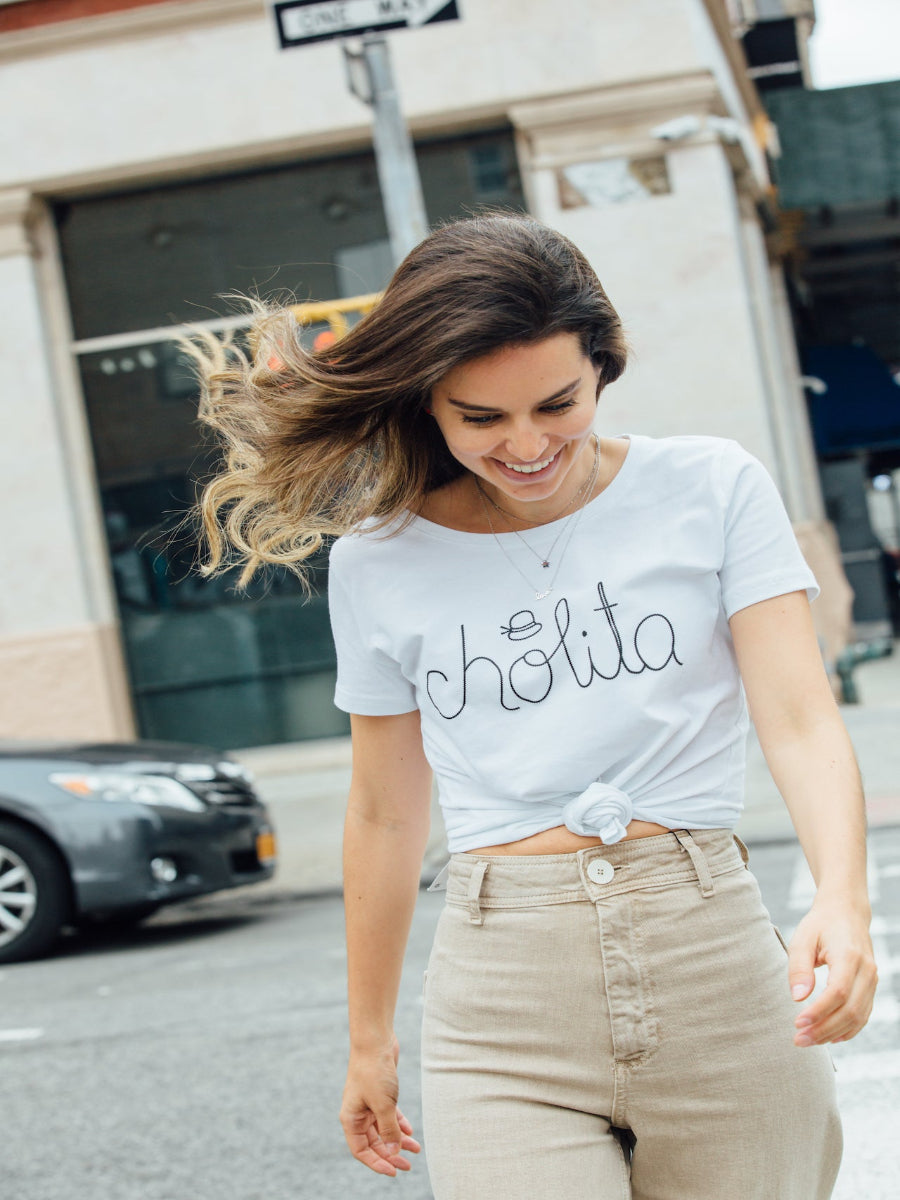 White cotton t-shirt featuring the word “cholita” embroidered in black script on the front, worn by a model in the street