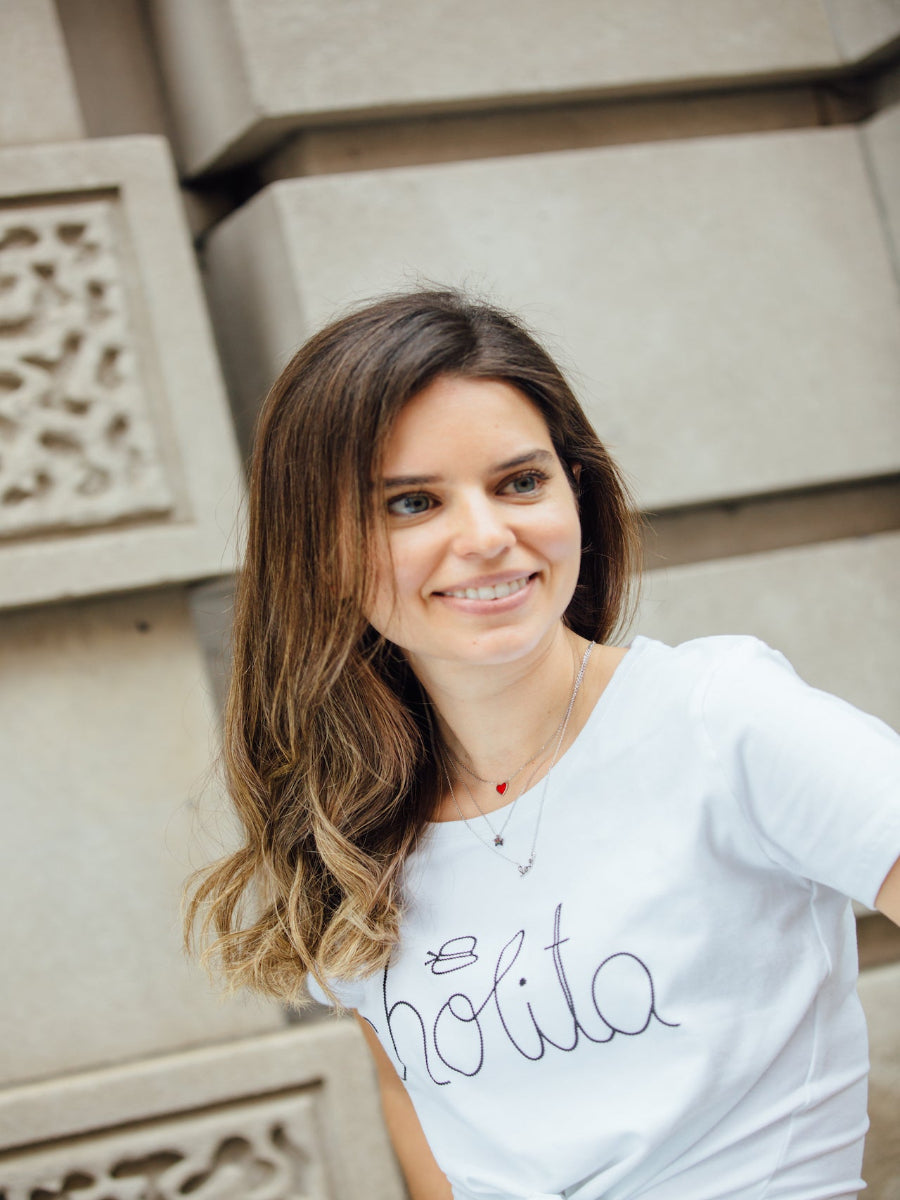 White cotton t-shirt featuring the word “cholita” embroidered in black script on the front, worn by a model in the street