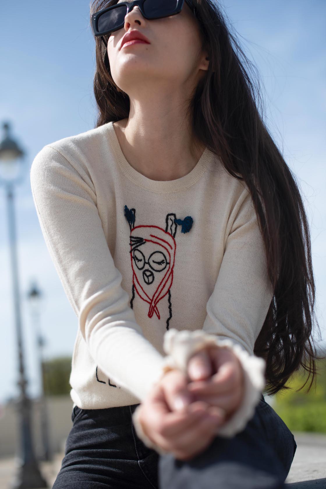 white alpaca sweater featuring a playful embroidered llama illustration with the word “LLAMOUR” on the front, shown here on a model in an outdoor setting.
