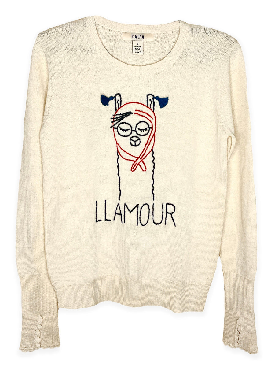 white alpaca sweater featuring a playful embroidered llama illustration with the word “LLAMOUR” on the front, shown here on a model in an outdoor setting.