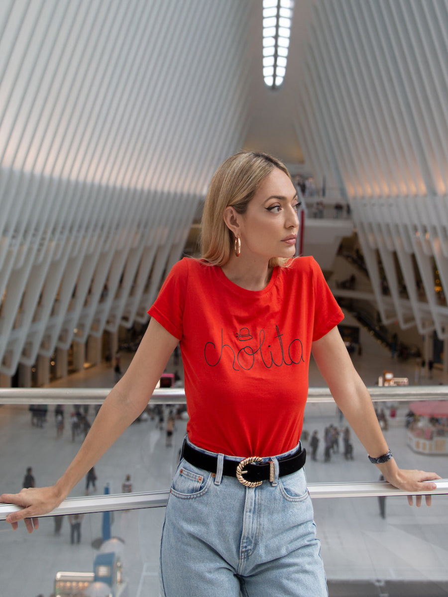 red cotton t-shirt featuring the word “cholita” embroidered in black script on the front, shown here on a model in an urban outdoor setting.
