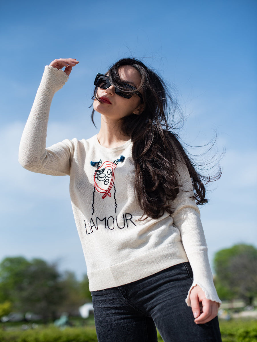 white alpaca sweater featuring a playful embroidered llama illustration with the word “LLAMOUR” on the front, shown here on a model in an outdoor setting.