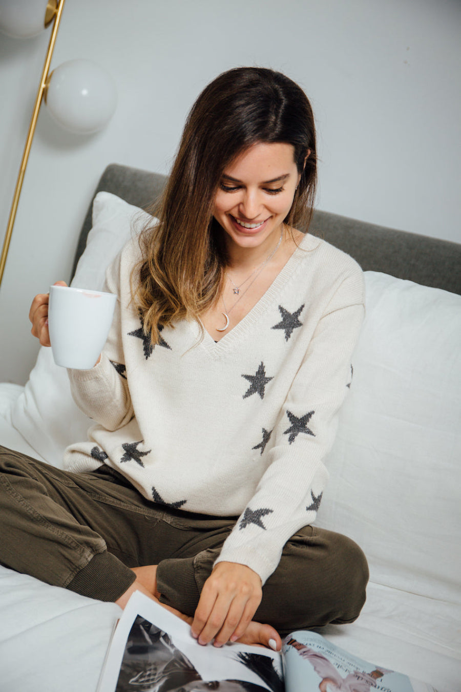 A white alpaca sweater with a V-neck and black star pattern, worn by a model sitting indoors on a bed